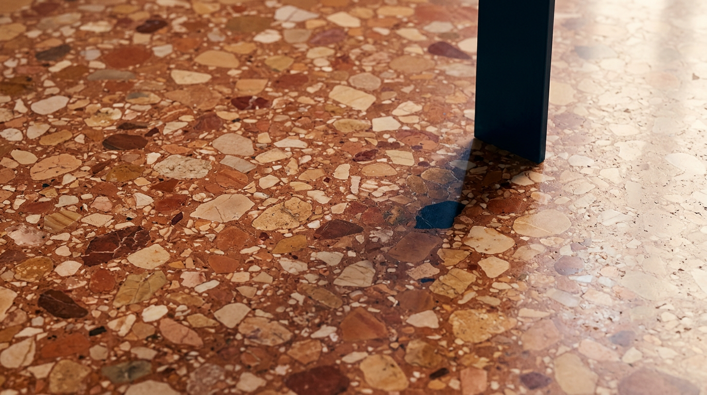 Close-up of a reddish-brown terrazzo floor with multicolored stone chips, partially shaded by a dark furniture leg.