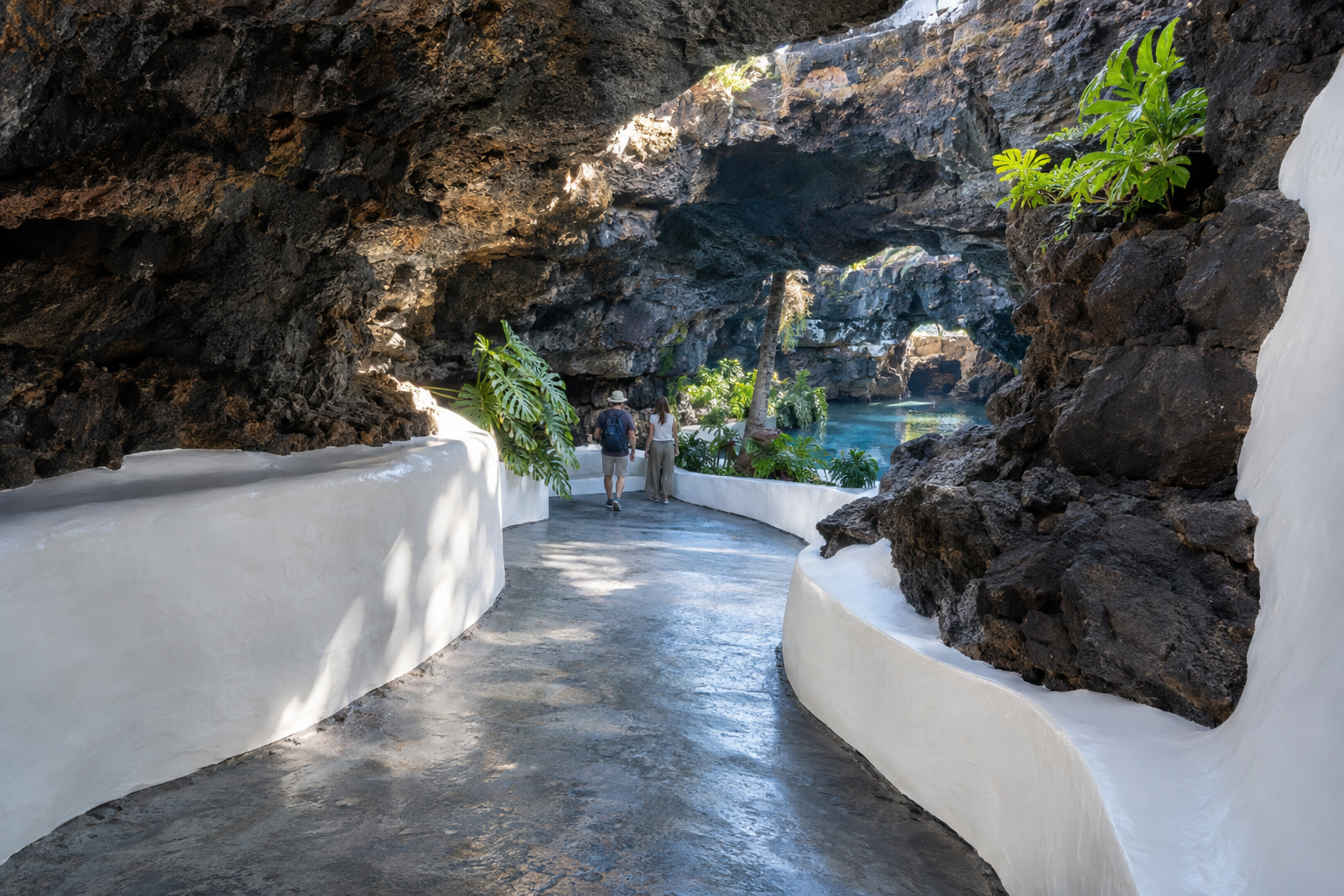 Curved white walls guide a concrete path through dark volcanic stone caves with a natural pool and plants.