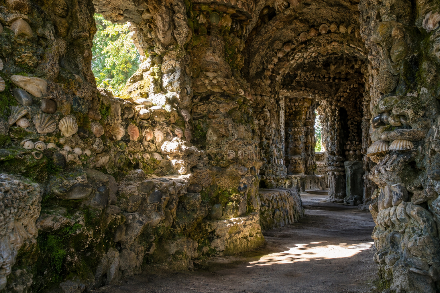 A grotto interior features rough stone walls and vaulted ceiling embedded with countless pebbles and seashells, with a window